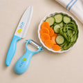 A children's kitchen knife and peeler set with light blue handles and white blades, styled on a beige surface next to a white bowl of sliced carrots and cucumbers.