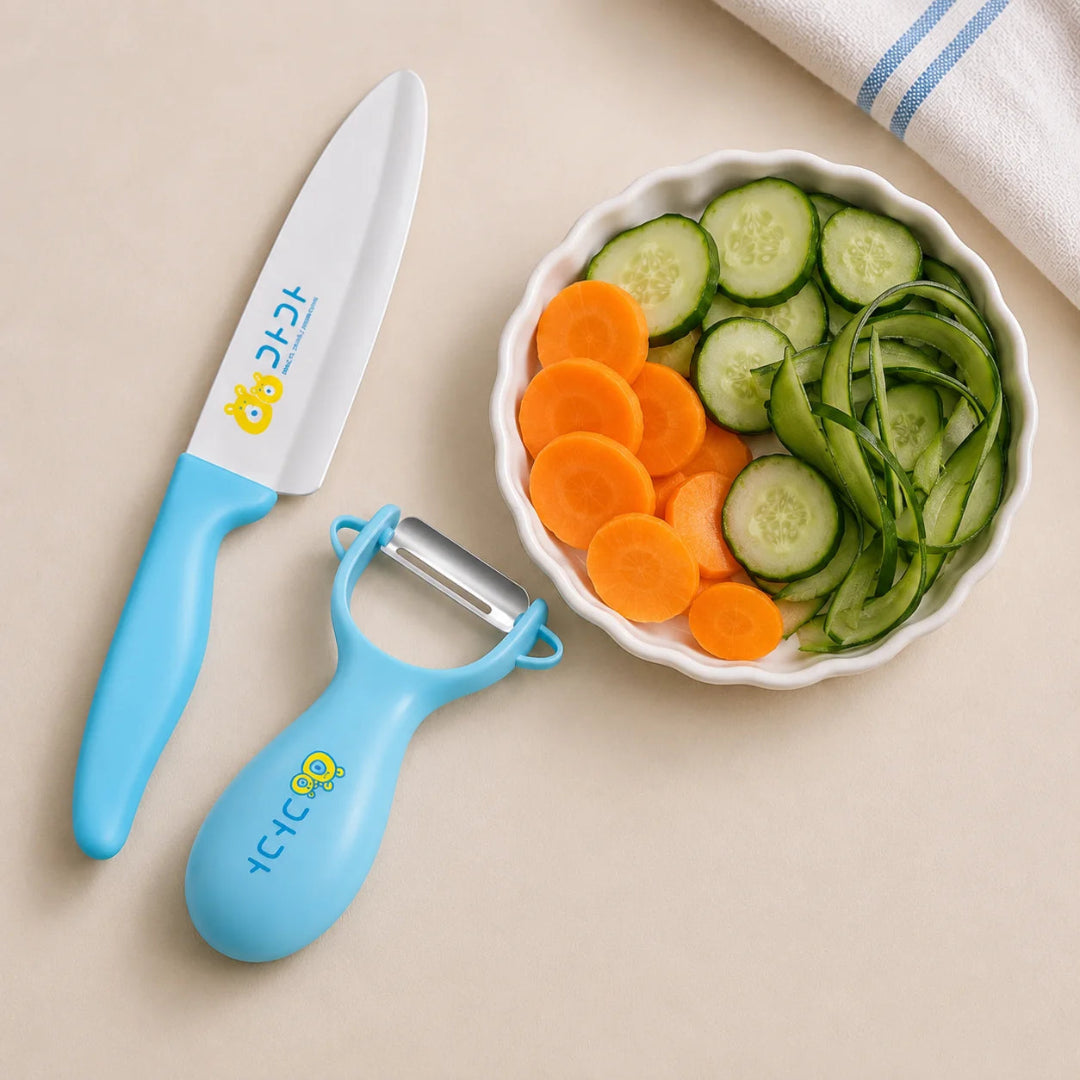 A children's kitchen knife and peeler set with light blue handles and white blades, styled on a beige surface next to a white bowl of sliced carrots and cucumbers.